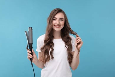 Happy young woman with beautiful hair holding curling iron on light blue background