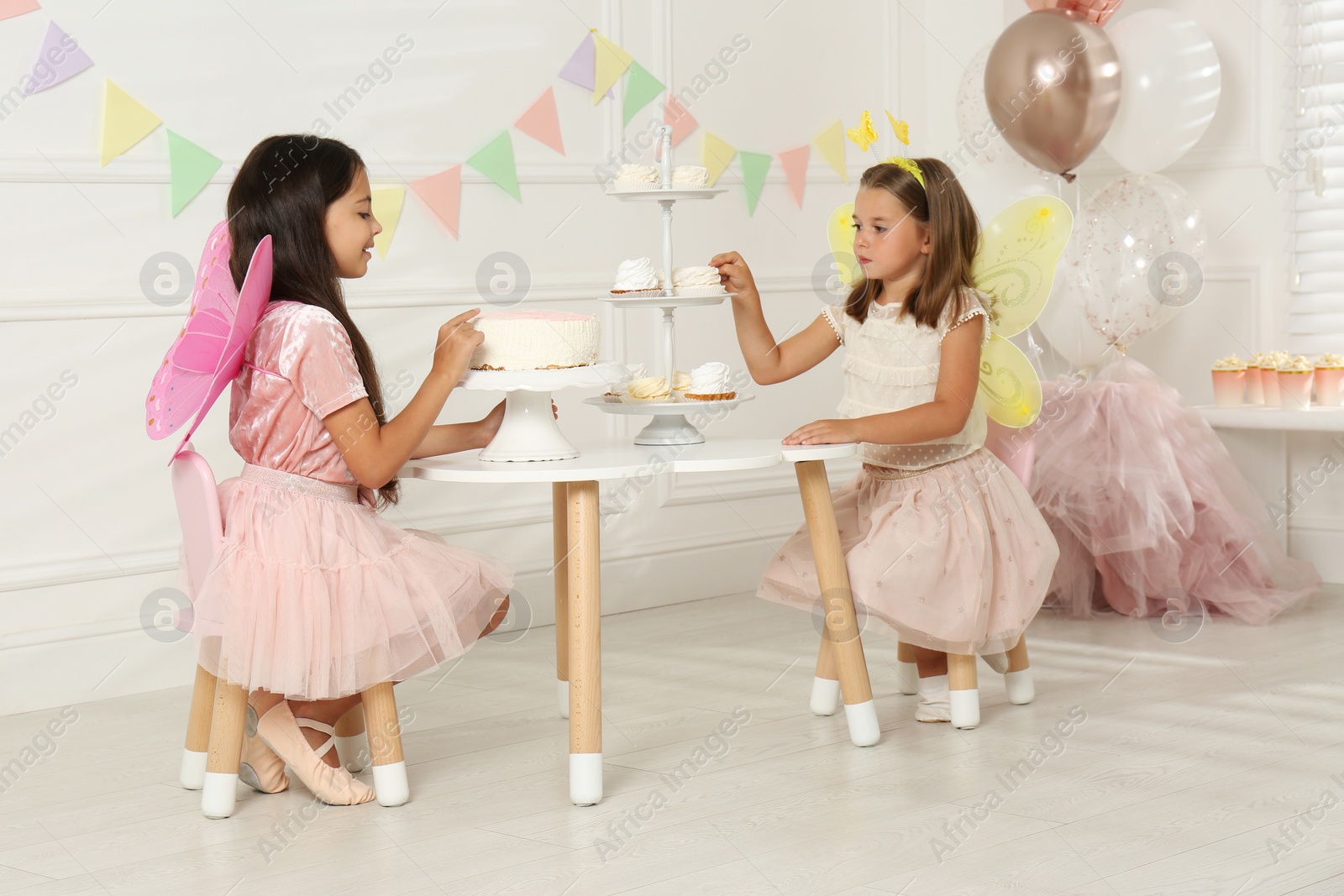 Photo of Cute little girls wearing fairy costumes at table with desserts in decorated room