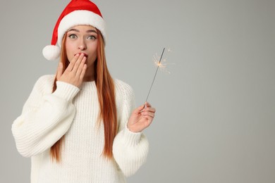 Photo of Emotional young woman in Santa hat with burning sparkler on light grey background, space for text. Christmas celebration