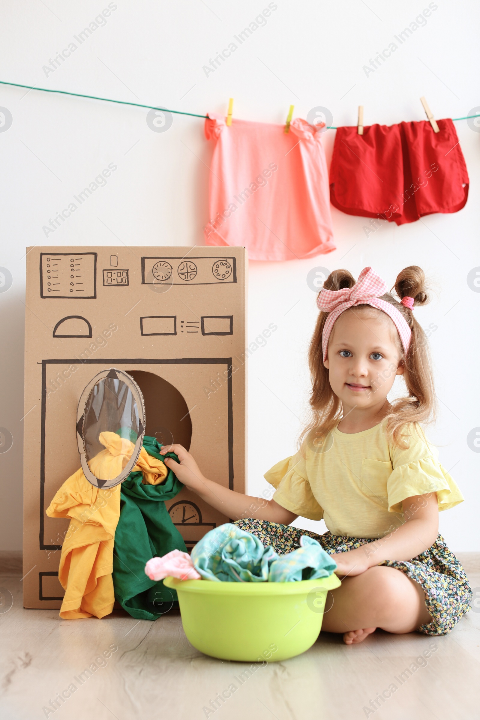 Photo of Adorable little child playing with cardboard washing machine and clothes indoors