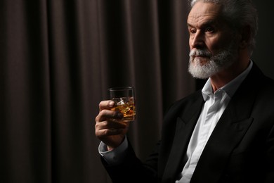 Photo of Senior man in suit holding glass of whiskey with ice cubes on brown background. Space for text