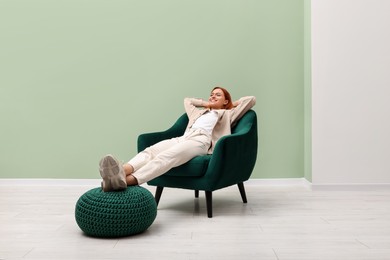 Happy young woman relaxing in armchair near light olive wall indoors