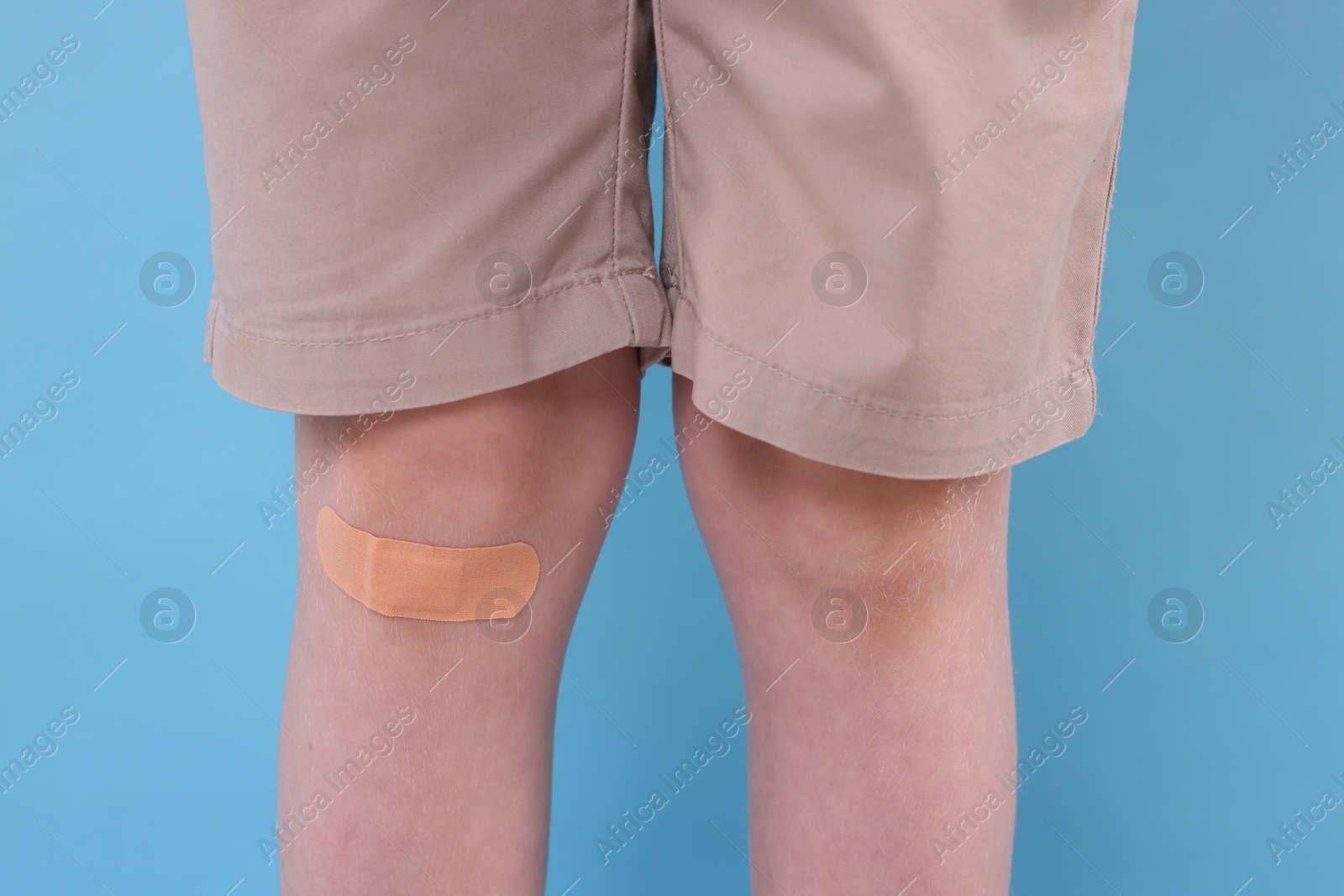 Photo of Little boy with sticking plaster on knee against light blue background, closeup