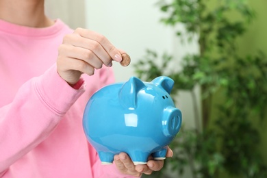 Woman putting coin into piggy bank indoors, closeup