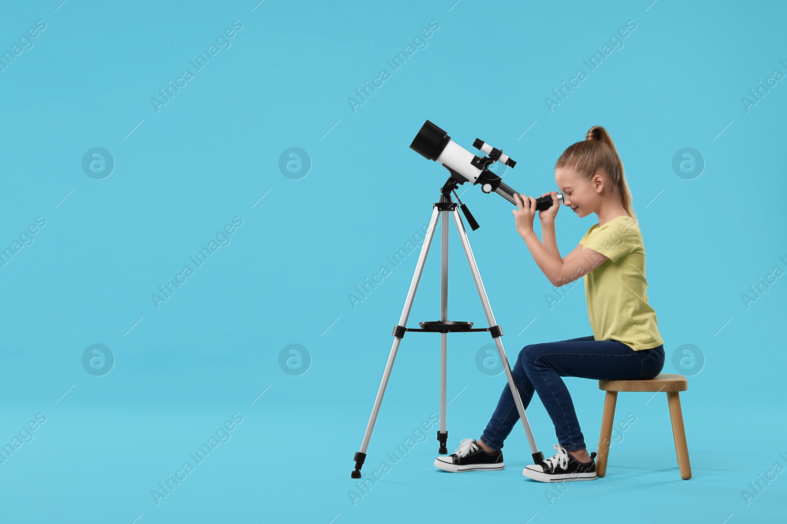 Photo of Happy girl looking at stars through telescope on light blue background, space for text