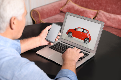 Photo of Man using laptop and phone to buy car at table indoors, closeup