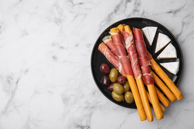 Photo of Plate of delicious grissini sticks with prosciutto, cheese and olives on white marble table, top view. Space for text