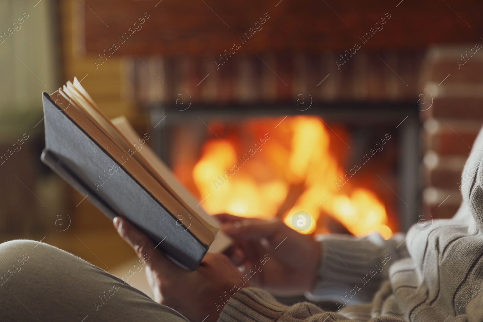 Photo of Man reading book near burning fireplace at home, closeup