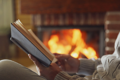 Photo of Man reading book near burning fireplace at home, closeup