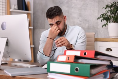 Photo of Overwhelmed man sitting at table with computer and stack of folders in office