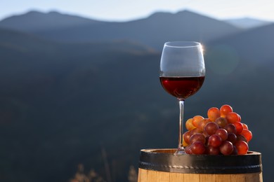 Photo of Glass of red wine and grapes on barrel against mountain landscape, space for text