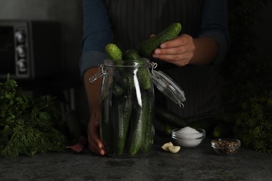 Woman putting cucumber into glass jar at black kitchen table, closeup. Pickling vegetables