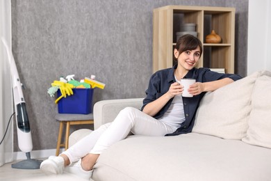 Beautiful young housewife with cup of drink resting after cleaning on sofa at home