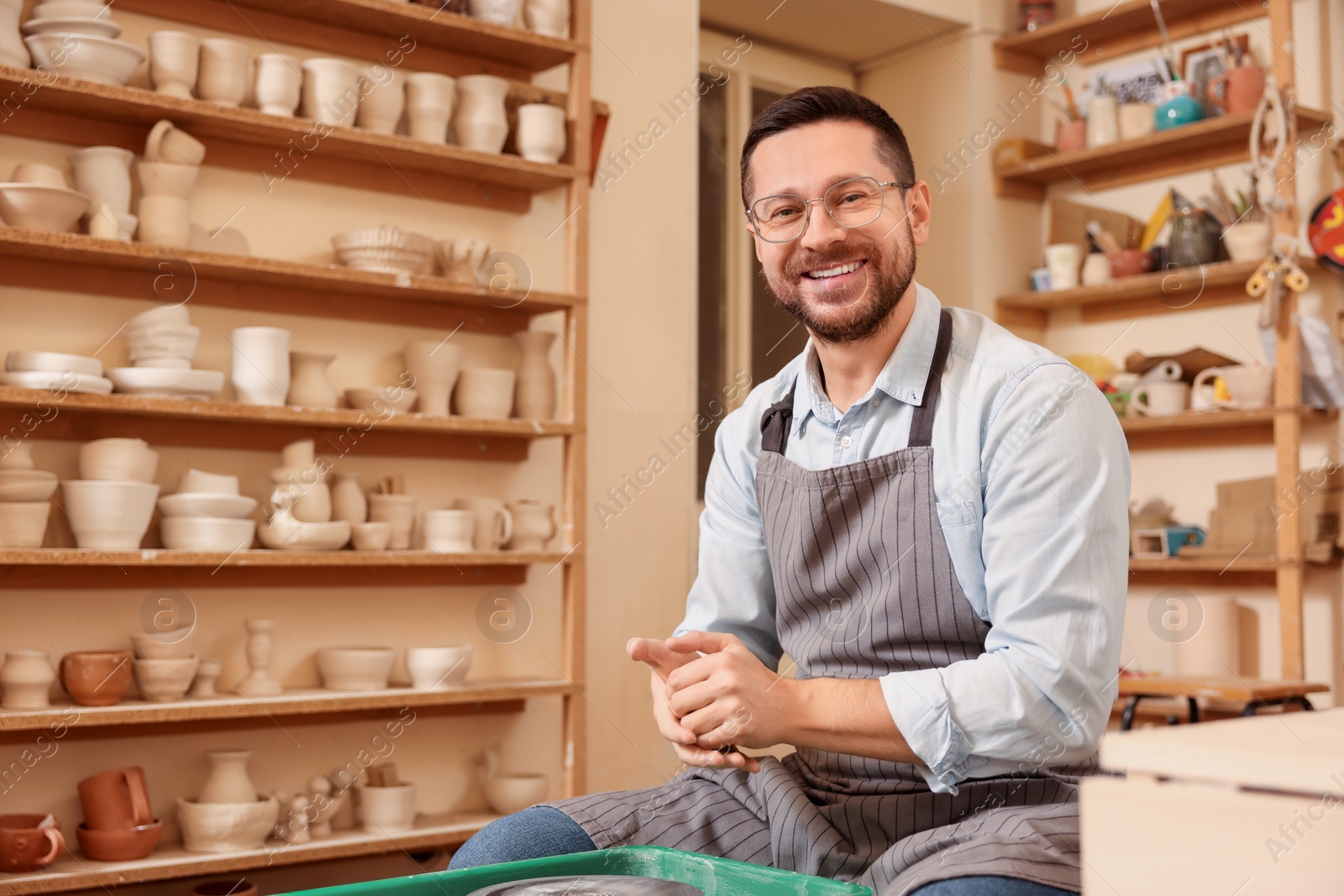 Photo of Happy man crafting with clay indoors. Different ceramics and potter's wheel in workshop