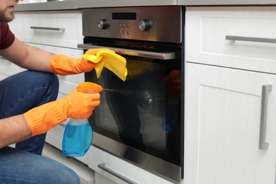 Young man cleaning oven with rag and detergent in kitchen, closeup