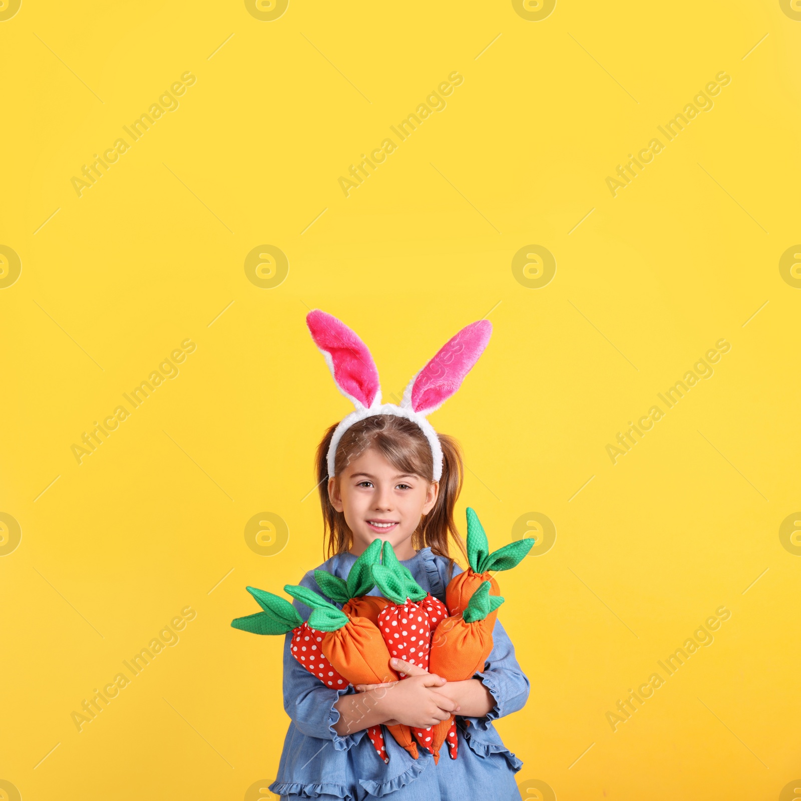 Photo of Happy little girl with bunny ears and handful of toy carrots on orange background. Easter celebration