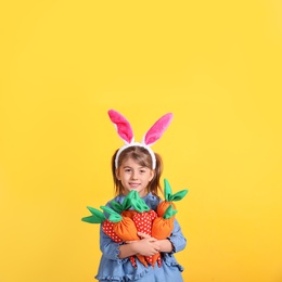 Photo of Happy little girl with bunny ears and handful of toy carrots on orange background. Easter celebration