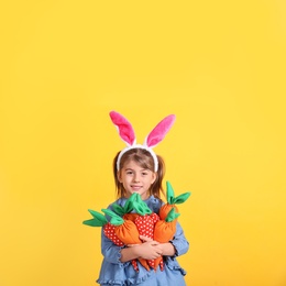 Happy little girl with bunny ears and handful of toy carrots on orange background. Easter celebration