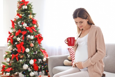 Photo of Happy pregnant woman with cup of tea sitting on sofa in room decorated for Christmas