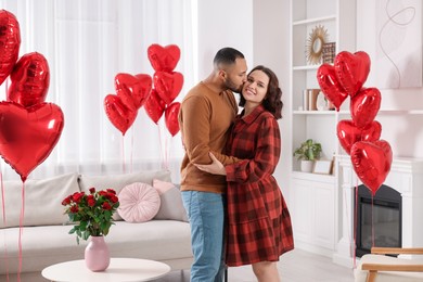 Photo of Man kissing his beloved woman in room decorated with heart shaped air balloons. Valentine's day celebration