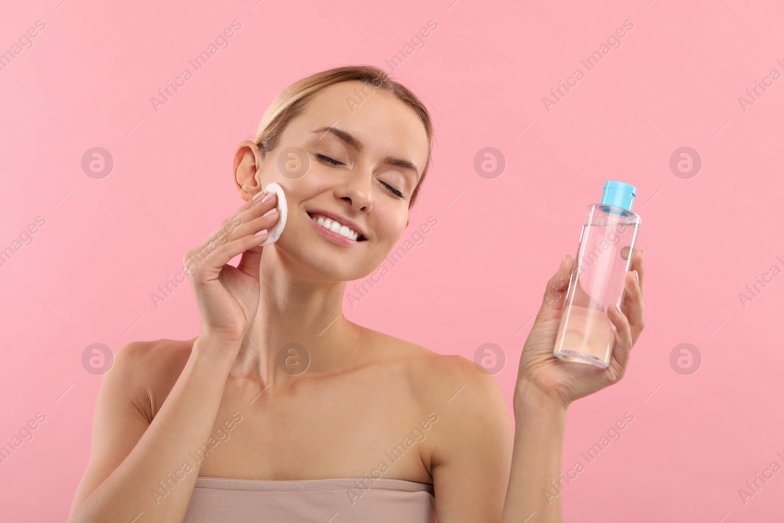 Photo of Smiling woman removing makeup with cotton pad and holding bottle on pink background