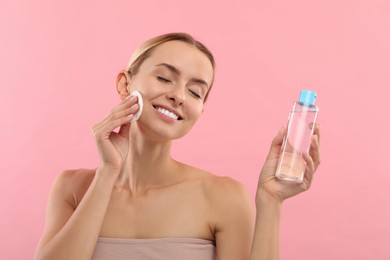 Smiling woman removing makeup with cotton pad and holding bottle on pink background