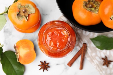 Photo of Jar of tasty persimmon jam and ingredients on white marble table, flat lay