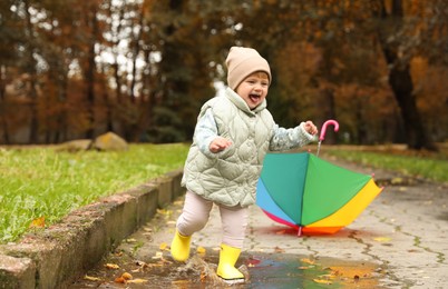 Cute little girl running in puddle near colorful umbrella outdoors