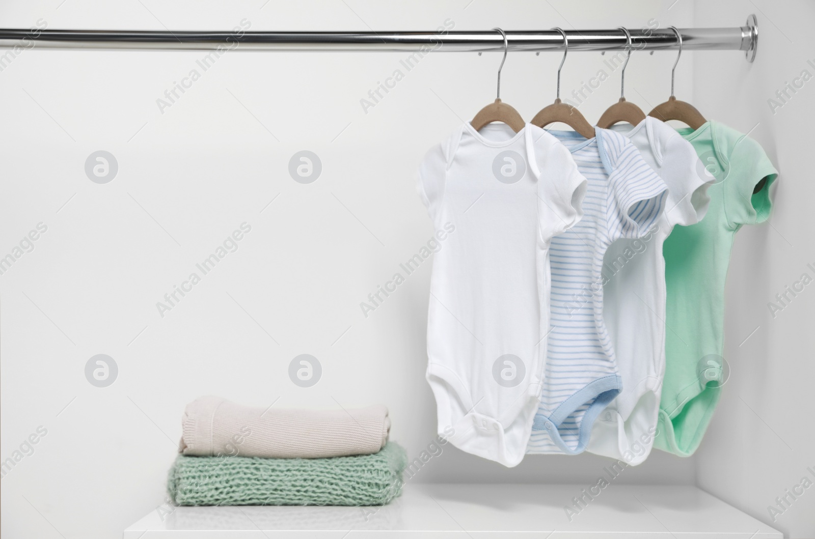 Photo of Hangers with baby bodysuits and stack of clothes near white wall. Space for text