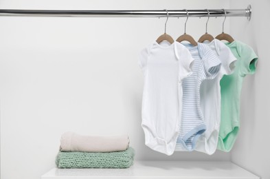 Hangers with baby bodysuits and stack of clothes near white wall. Space for text