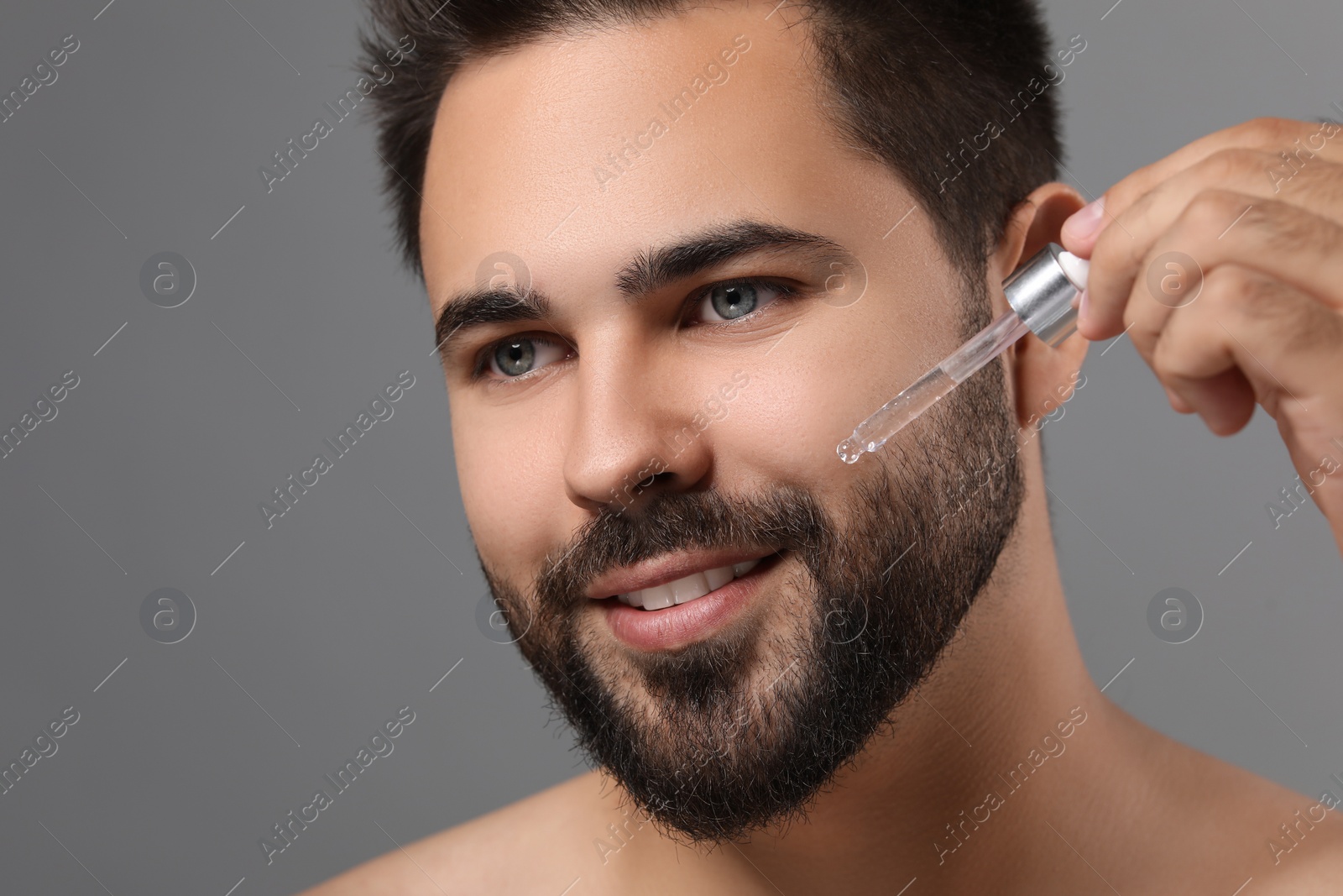 Photo of Handsome man applying cosmetic serum onto face on grey background, closeup