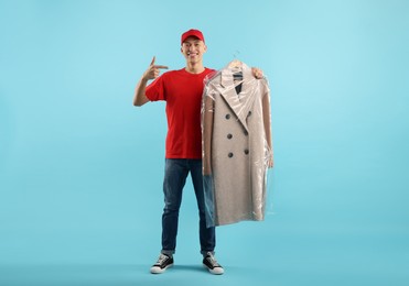 Dry-cleaning delivery. Happy courier holding coat in plastic bag on light blue background