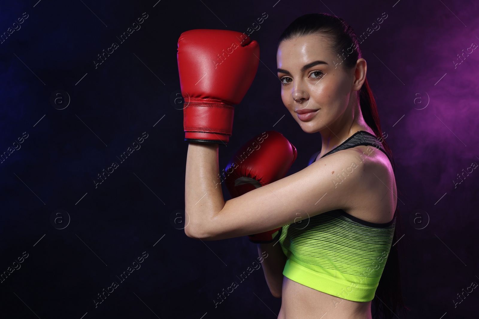 Photo of Portrait of beautiful woman wearing boxing gloves training in color lights and smoke on black background. Space for text