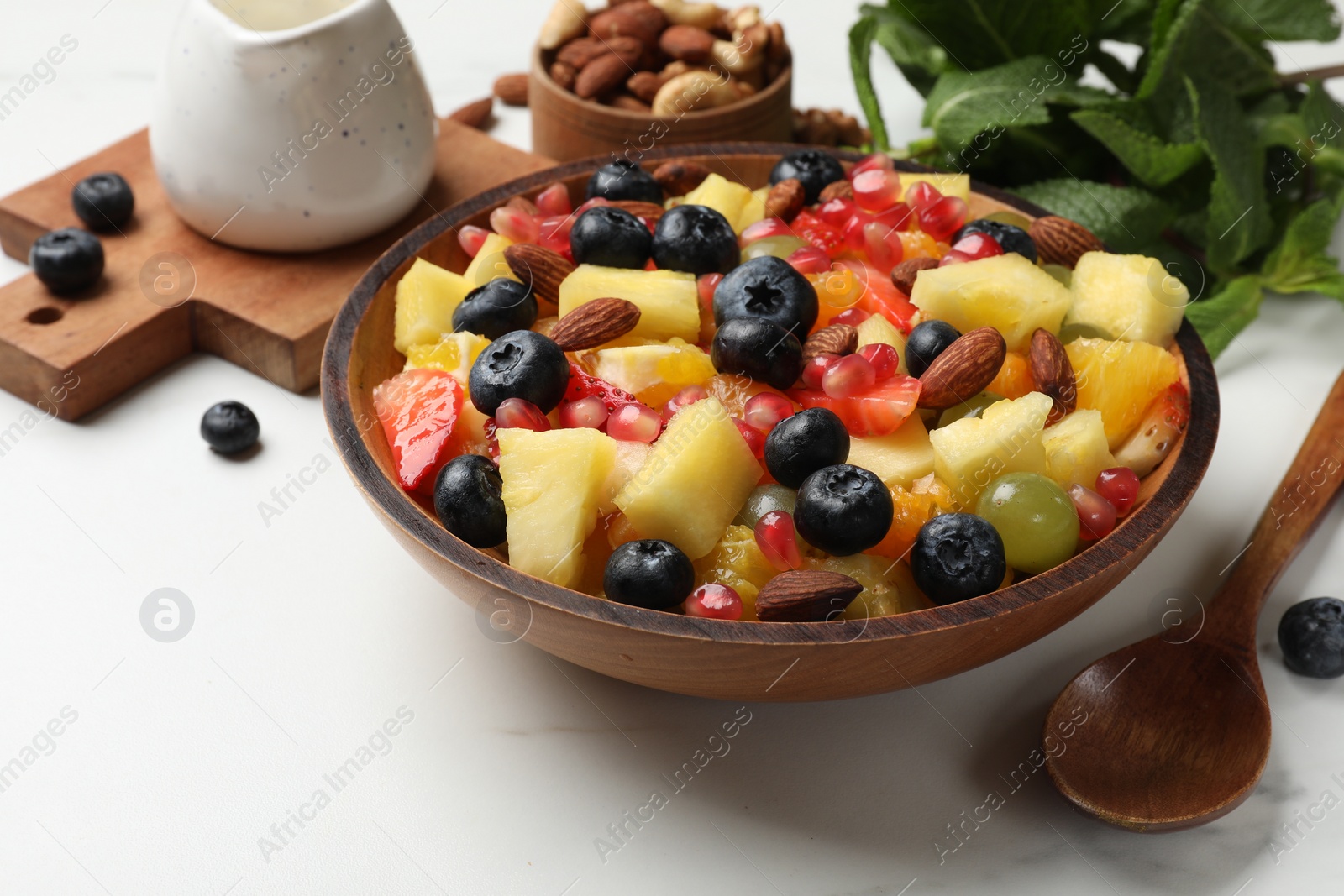 Photo of Delicious fruit salad in bowl, berries, fresh mint and nuts on white marble table