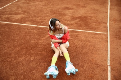Happy stylish young woman with vintage roller skates and headphones sitting on tennis court