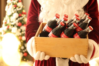 MYKOLAIV, UKRAINE - JANUARY 18, 2021: Santa Claus holding wooden crate full of Coca-Cola bottles in room with Christmas tree, closeup