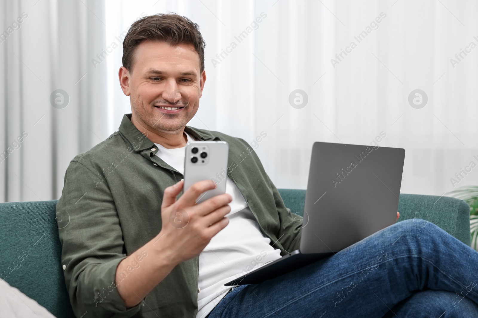 Photo of Happy man with laptop using smartphone on sofa at home