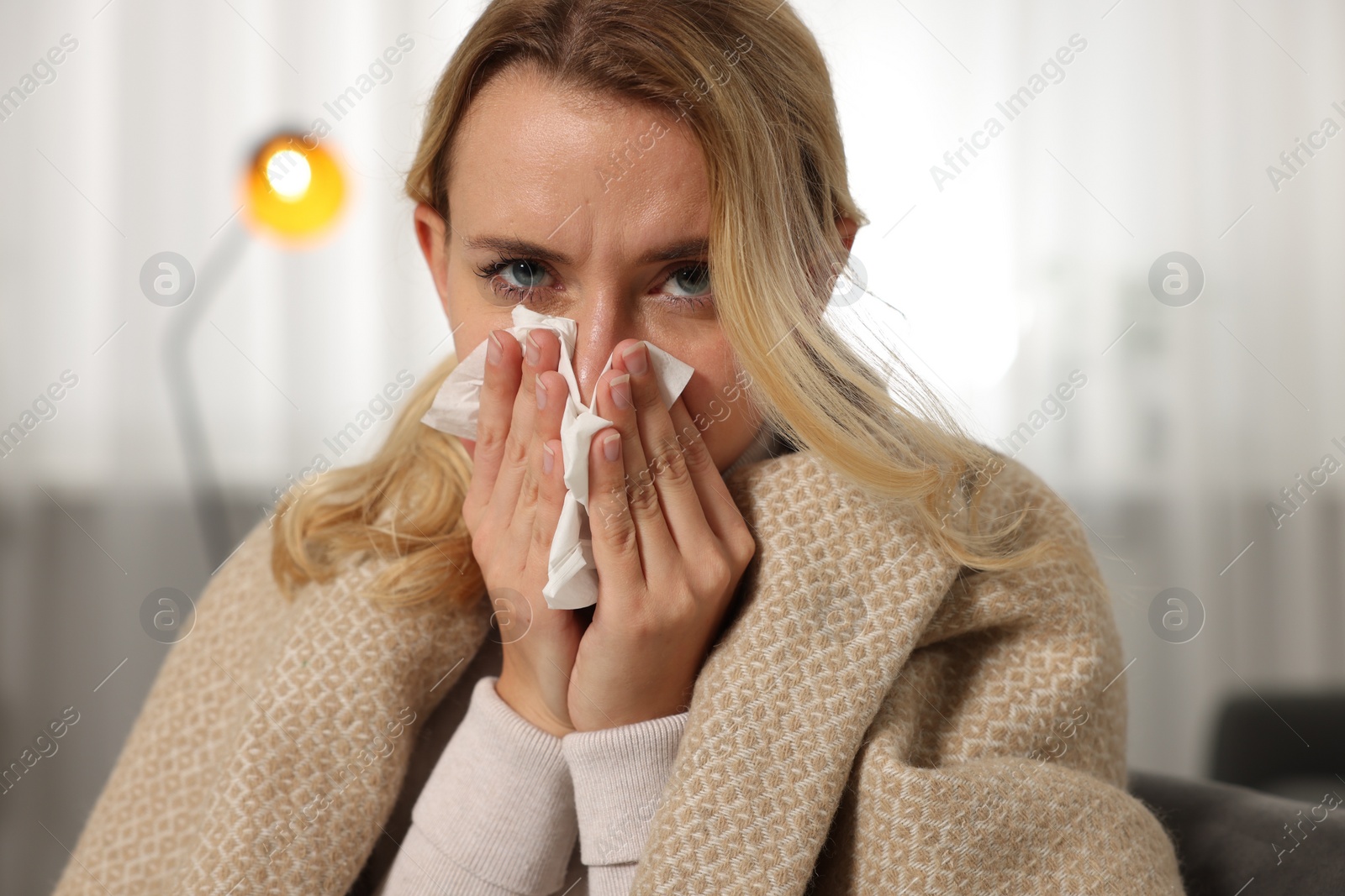 Photo of Sick woman wrapped in blanket blowing nose in tissue at home. Cold symptoms