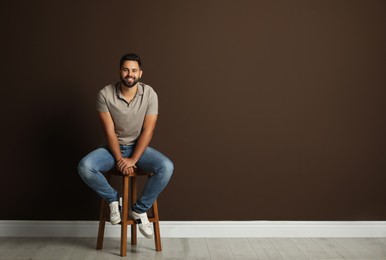 Handsome young man sitting on stool near brown wall. Space for text