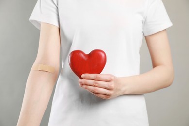 Photo of Blood donation concept. Woman with adhesive plaster on arm holding red heart against grey background, closeup