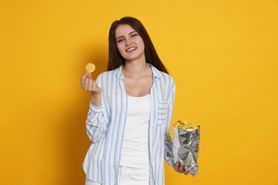 Pretty young woman with bag of tasty potato chips on yellow background