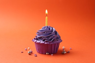 Photo of Delicious birthday cupcake with burning candle and sprinkles on coral background