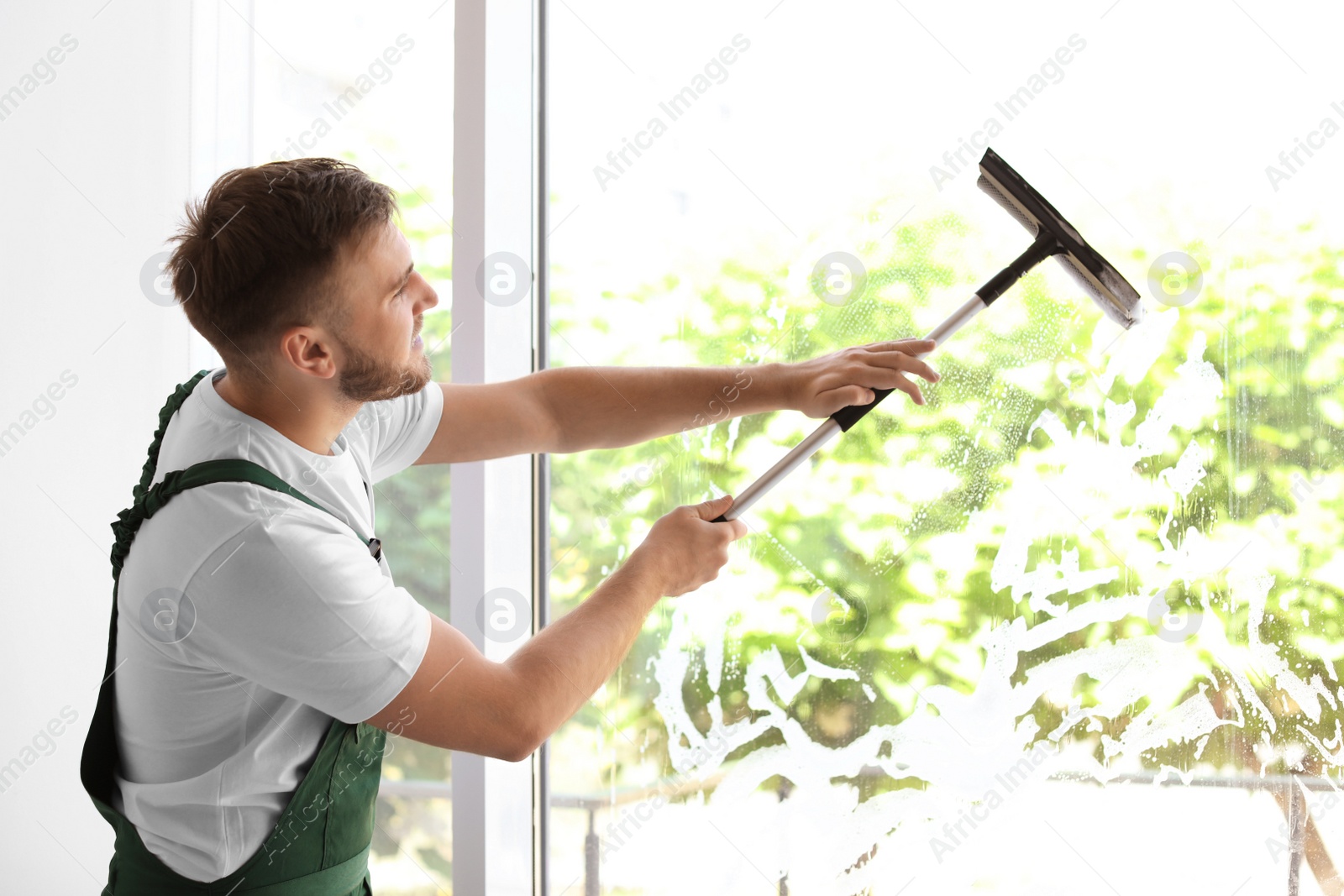 Photo of Male cleaner wiping window glass with squeegee indoors