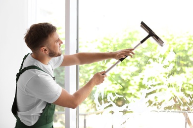 Male cleaner wiping window glass with squeegee indoors