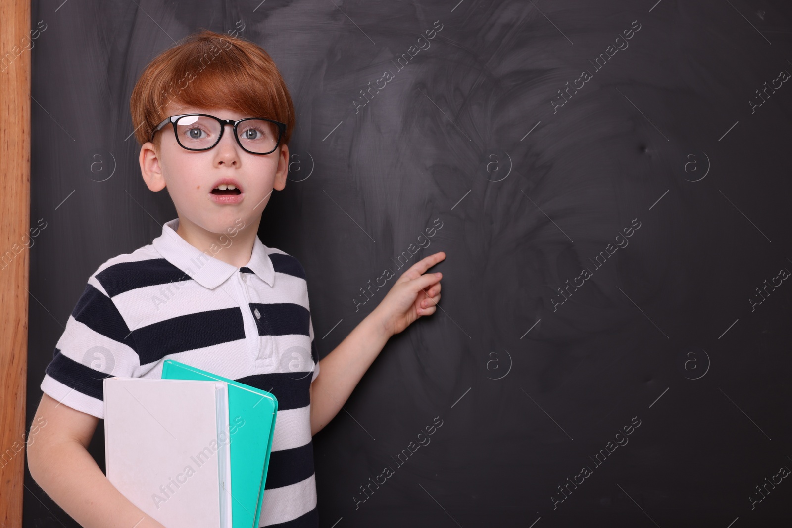 Photo of Shocked schoolboy in glasses with books pointing at something on blackboard. Space for text