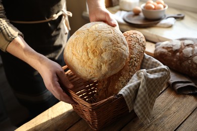 Photo of Man holding wicker basket with different types of bread at wooden table indoors, closeup