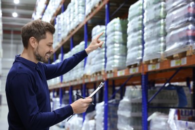 Happy manager holding clipboard in warehouse with lots of products. Space for text