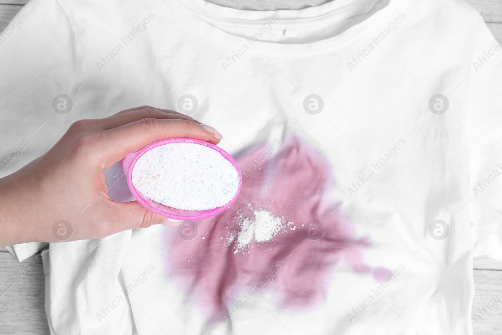 Photo of Woman adding powdered detergent onto white t-shirt with stain on wooden surface, top view. Hand washing laundry