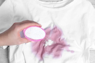 Photo of Woman adding powdered detergent onto white t-shirt with stain on wooden surface, top view. Hand washing laundry