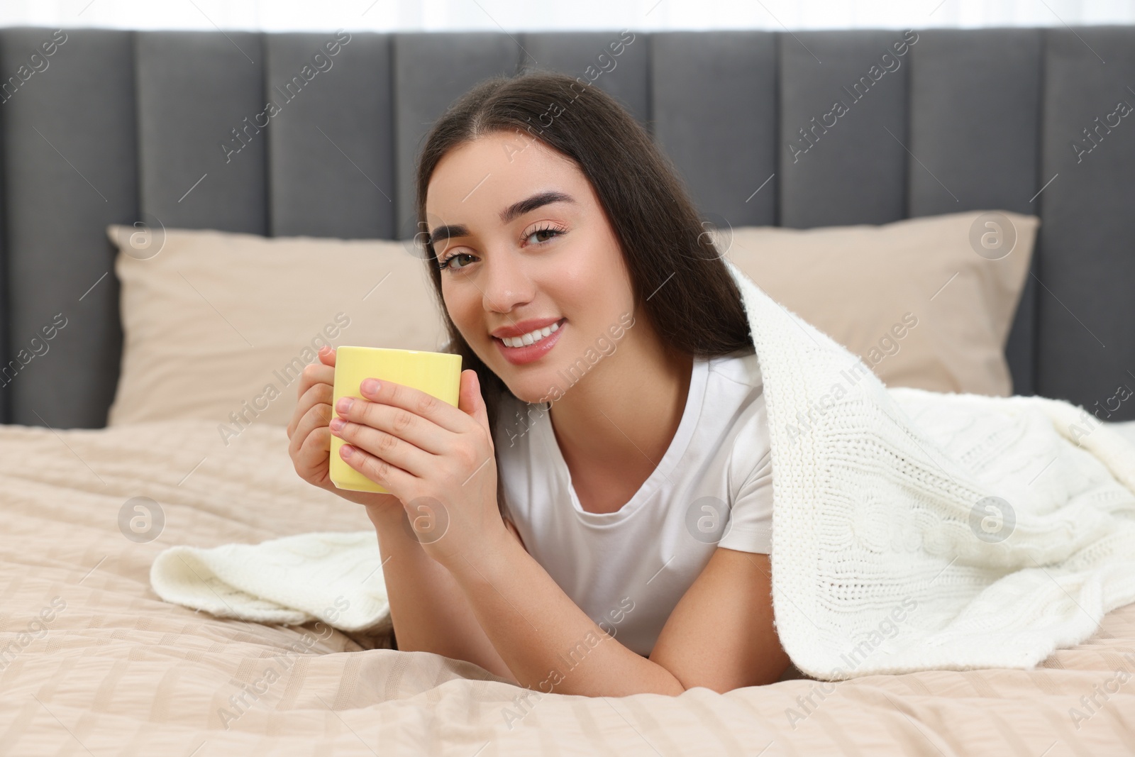 Photo of Happy young woman under plaid holding yellow ceramic mug on bed